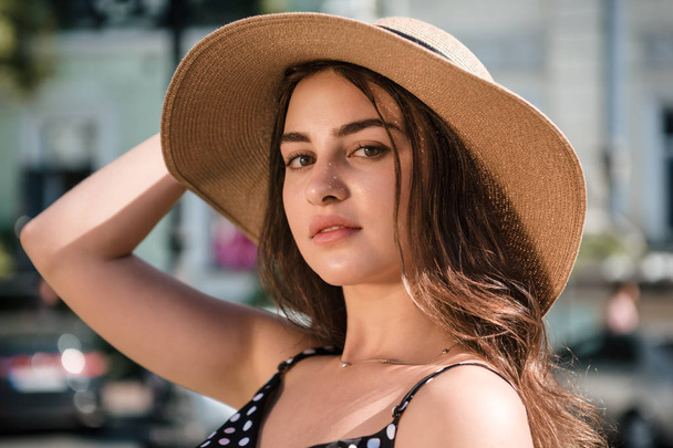 Bella ragazza in un abito nero e un cappello con ampi risvolti sorride godendo camminare lungo la città europea durante la soleggiata giornata estiva
. - Foto, immagini