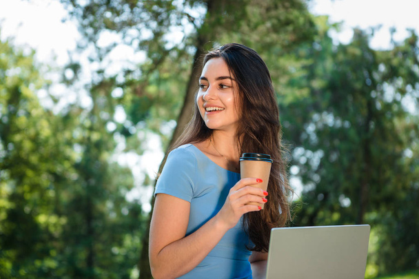 Νεαρή μελαχρινή ελεύθερος επαγγελματίας σε ένα μπλε φόρεμα που κάθεται με ένα φορητό υπολογιστή σε ένα πάρκο και πίνοντας καφέ από ένα χάρτινο κύπελλο μίας χρήσης. Εργαστείτε σε οποιοδήποτε μέρος - Φωτογραφία, εικόνα