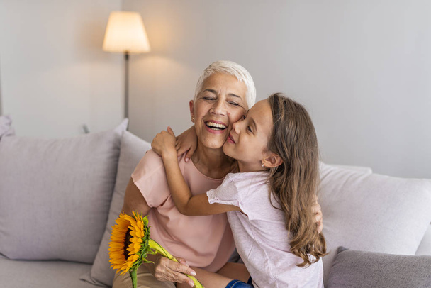 Cute girl giving a bunch of flowers to her grandmother. Happy senior grandma hugging granddaughter thanking for gift and flowers. Little granddaughter kissing giving flowers bouquet congratulating smiling old grandmother - Photo, image