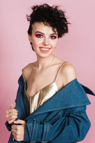 Een meisje met een oprechte brede glimlach staat in de studio op een pastel roze achtergrond. Een meisje met een vreemd vliegende kapsel en heldere make-up. - Foto, afbeelding