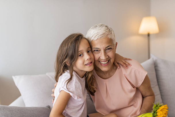 Granddaughter embracing her grandmother in living room. Lovely little girl with her grandmother looking at the camera. Girl and grandma on a sofa Smiling senior woman and girl embracing - Foto, Bild