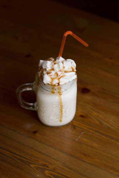 Milk shake in glass jar with whipped cream, marshmallow, pumpkin and caramel syrup, orange straw on wooden table, autumn fall October mood, with yellow pillow on background. Dark food beverage photo - Photo, Image