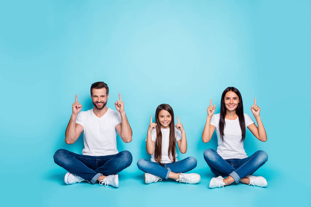 Full length body size photo of fascinating cheerful charming casual family pointing up for you to see something sitting with legs crossed while isolated with blue background with copyspace - Foto, imagen