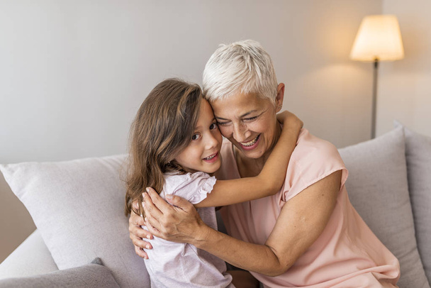 Cherishing her grandmother. Granddaughter embracing her grandmother in living room. Lovely little girl with her grandmother Girl and grandma on a sofa  - Foto, afbeelding