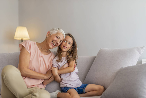 Granddaughter embracing her grandmother in living room. Grandmother's give the best hugs. Girl and grandma on a sofa. Smiling senior woman and girl embracing - 写真・画像
