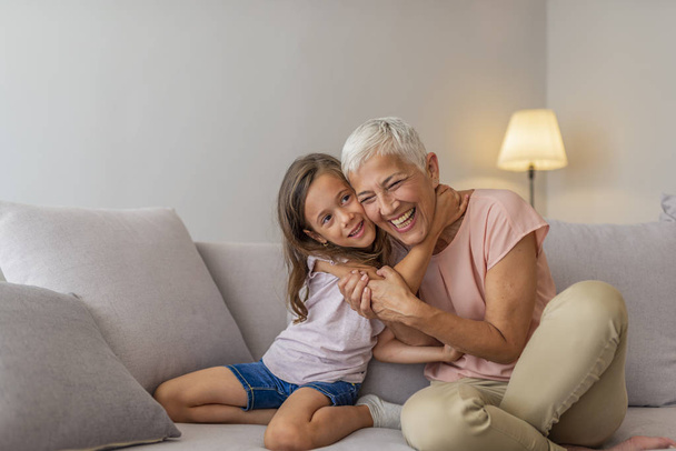 Granddaughter embracing her grandmother in living room. Grandmother's give the best hugs. Girl and grandma on a sofa. Smiling senior woman and girl embracing - Photo, image