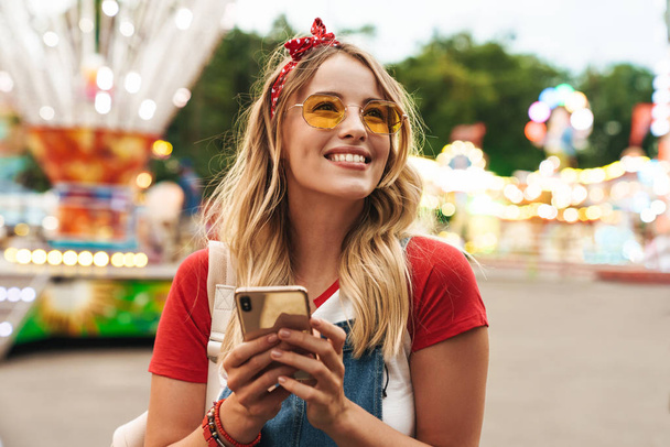 Image of cute blonde woman holding cellphone and walking in fron - Foto, afbeelding