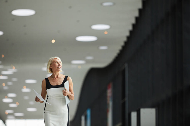Зрелая улыбающаяся деловая женщина в элегантном платье с ноутбуком и документами и идущая по офисному коридору  - Фото, изображение