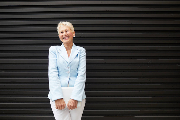 Portrait of mature successful businesswoman with laptop standing outdoors in the city and laughing - Photo, Image