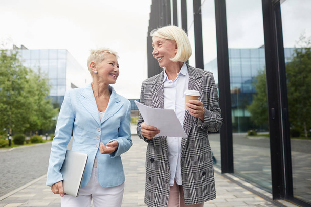 Two mature businesswomen with laptop and documents walking along the street drinking coffee and talking   - 写真・画像