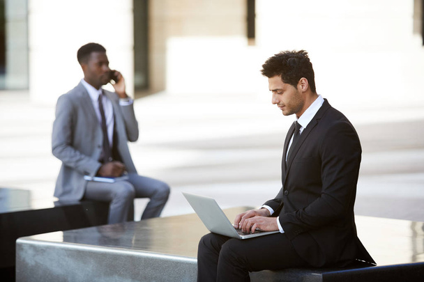 Pensive бізнесмен сидить з ноутбуком на колінах, працює на відкритому повітрі в місті з людиною на задньому плані
 - Фото, зображення