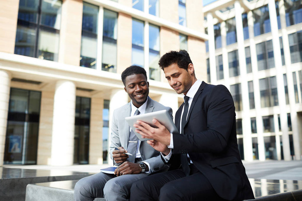 Kaksi nuorta liikemiestä puvuissa katsomassa online-konferenssia digitaalisella tabletilla ja hymyilemässä istuessaan ulkona kaupungissa toimistorakennus taustalla - Valokuva, kuva