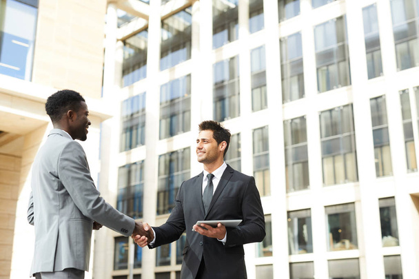 Zwei Geschäftspartner schütteln Hände und grüßen im Freien mit Bürogebäude im Hintergrund - Foto, Bild