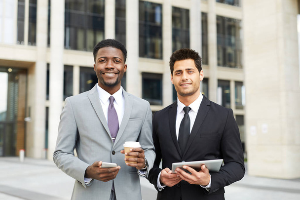 Портрет двух молодых менеджеров в формальной одежде, стоящих с мобильным телефоном и планшетным ПК и улыбающихся в камеру с современным зданием на заднем плане - Фото, изображение