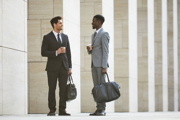 Δύο πολυεθνικοί επιχειρηματίες με κοστούμια κρατούν τσάντες πίνοντας καφέ και μιλώντας μεταξύ τους ενώ στέκονται στην πόλη - Φωτογραφία, εικόνα