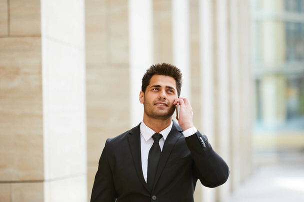 Junger Geschäftsmann im schwarzen Anzug mit Tasche, der auf der Straße in der Stadt mit dem Handy telefoniert  - Foto, Bild