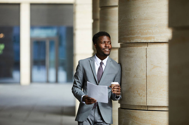 Молодий африканський бізнесмен у костюмі, який спирається на колонку офісних будівель, читає документ і п "є каву просто неба. - Фото, зображення