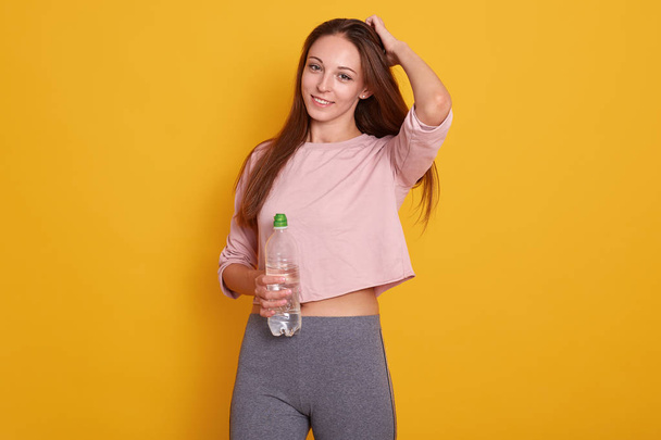 Studio shot of caucasian fitness woman model standing in sporty gym clothing, having dark straight hair, holds water bottle, posing isolated over yellow background. Sport, healthy lifestyle concept. - Valokuva, kuva