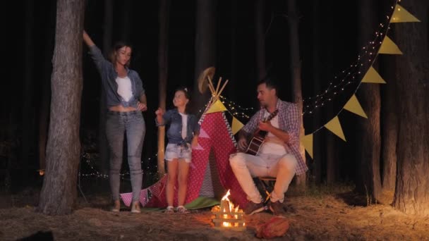 Unbekümmerte Kindheit, schönes Mädchen, das mit ihrer liebenden Mutter zur Musik ihres Vaters tanzt, der beim nächtlichen Picknick in der Waldkulisse von Wigwam Gitarre spielt - Filmmaterial, Video