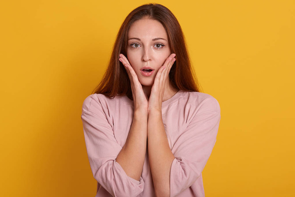 Close up portrait of emotional female being shocked to see something, opens mouth with surprisment and keepspalms near cheek, poses against yellow studio background. People emotions concept. - Photo, Image