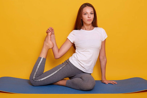 Horizontal shot of slim sporty woman doing stretching exercises in gym, female sitting on yogamat on floor, dresses whitet shirt and leggins, looking directrly at camera. Healthy lifestyle concept. - Фото, зображення