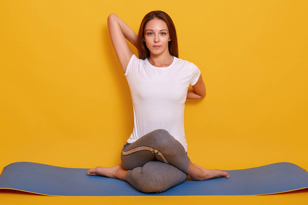 Horizontal shot of flexible girl doing yoga pose isolated over yellow background, adorable female sitting on floor on yoga mat, looking directly at camera. sport and healthy lifestyle concept. - Фото, зображення