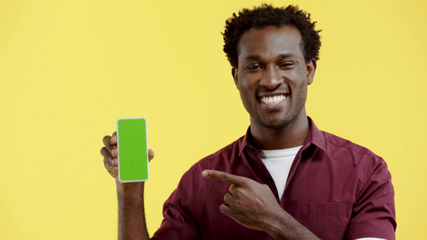 glimlachende Afrikaans-Amerikaanse man wijzend naar smartphone geïsoleerd op geel - Video