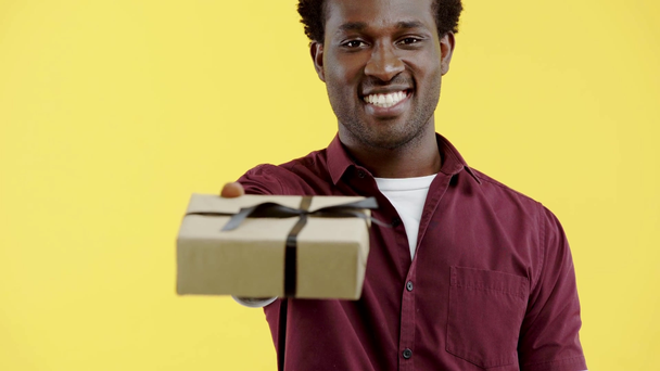όμορφος Αφροαμερικάνος παρουσιάζοντας κουτί δώρου απομονωμένο στο κίτρινο - Πλάνα, βίντεο
