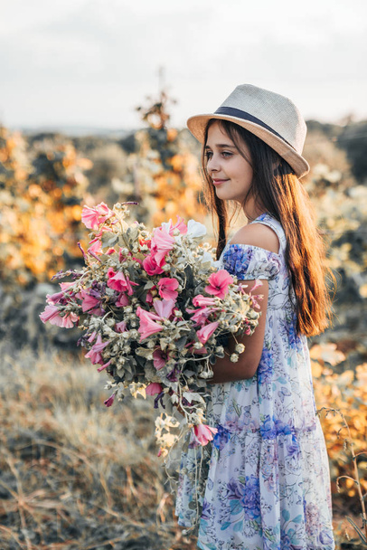 Side view portrait of little cute brunette girl in dress with flowers and white hat with a large beautiful bouquet of pink flowers - Фото, изображение