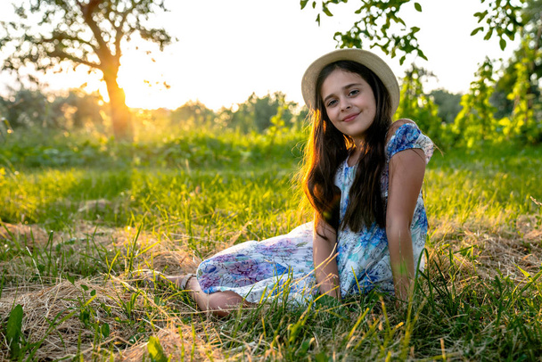Menina bonita feliz com cabelo morena vestindo vestido com flores imprimir e chapéu branco sentado no prado com grama seca cortada e olhando para a câmera, pôr do sol no fundo
 - Foto, Imagem