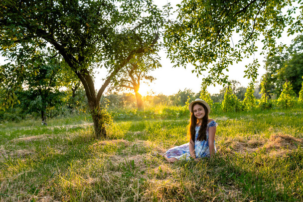 Happy smiling teen girl in white hat and dress with flowers print sitting on the meadow among the trees and looking at the camera, sunset on the background - Photo, Image