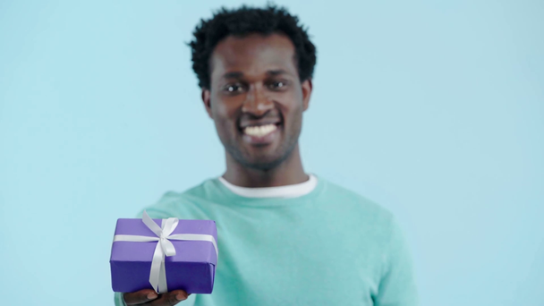 χαμογελαστός Αφροαμερικάνος που κρατάει κουτί δώρου απομονωμένο στο μπλε - Πλάνα, βίντεο