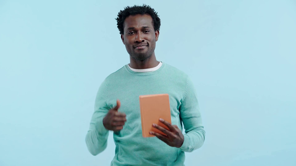 happy african american man reading book and showing thumb up isolated on blue - Кадры, видео