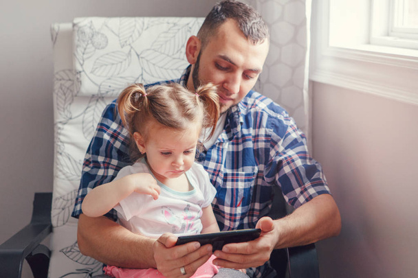 Caucasian bearded father dad sitting with daughter girl child watching cartoons on smartphone digital tablet together. Candid lifestyle family life. Single parent babysitting kid at home. - Zdjęcie, obraz