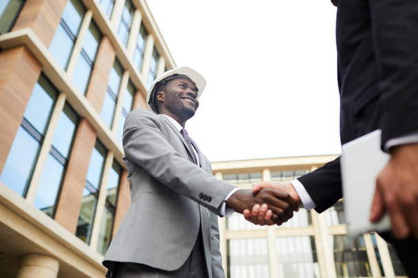 African young engineer in suit and hardhat shaking hands to partner and smiling while standing in city - Photo, Image