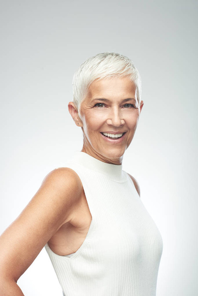 Beautiful smiling senior woman with short gray hair posing in front of gray background. Beauty photography. - Foto, afbeelding
