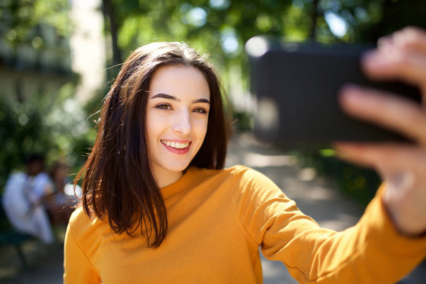 Портрет красивой молодой женщины, делающей селфи с мобильного телефона в парке
 - Фото, изображение