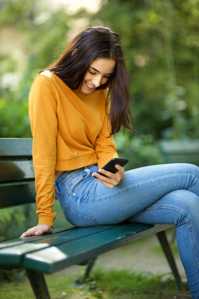 Porträt einer jungen Frau, die auf einer Parkbank sitzt und auf ihr Handy schaut - Foto, Bild