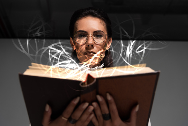 niski kąt widzenia zamyślony steampunk kobieta w okularach czytania książki z świecącą ilustracją powyżej - Zdjęcie, obraz