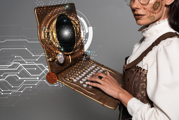καλλιεργημένη άποψη της γυναίκας steampunk χρησιμοποιώντας vintage laptop με λαμπερό ψηφιακή απεικόνιση απομονώνονται σε γκρι - Φωτογραφία, εικόνα