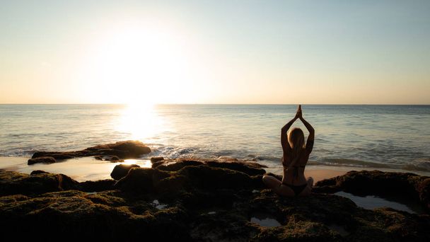 Woman sitting on the rock, practicing yoga and enjoying ocean view. View from back. Hands in namaste mudra. Yoga on the beach, Bali - Photo, Image