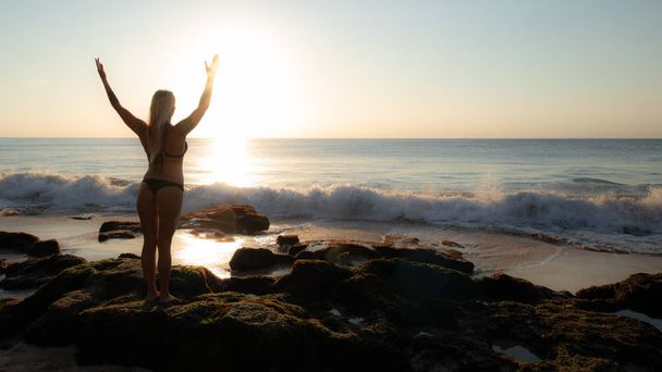 Excited young woman raising arms at the beach in front of the ocean. View from back. Sunset at the beach. Bali, Indonesia. - Foto, Imagen