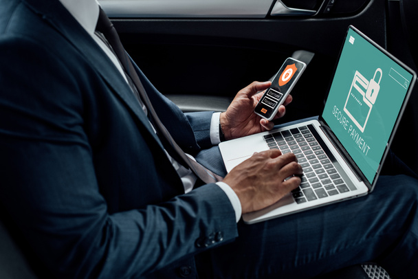 vue recadrée d'un homme d'affaires afro-américain utilisant un ordinateur portable et un smartphone dans une voiture avec une illustration de cybersécurité
 - Photo, image