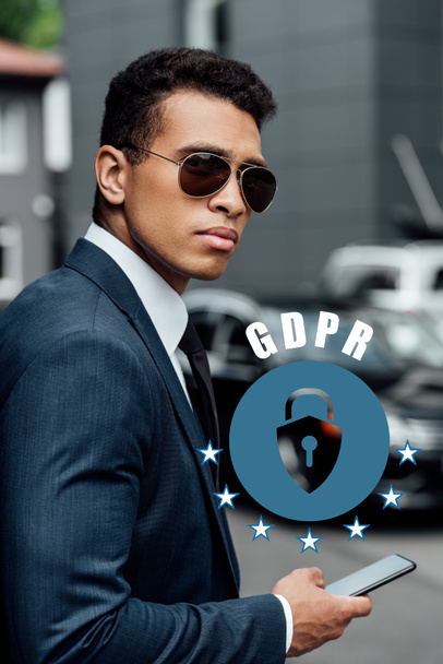 hombre de negocios afroamericano guapo y confiado en traje y gafas de sol usando teléfono inteligente con ilustración gdpr
 - Foto, Imagen