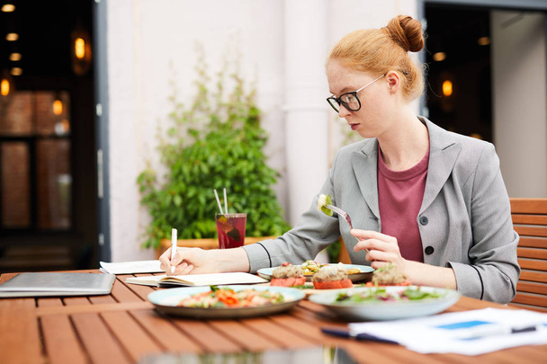 Primer plano de la joven sentada a la mesa y comiendo ensalada de verduras durante el almuerzo de negocios en la cafetería al aire libre
 - Foto, imagen