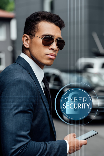 Yakışıklı ve kendinden emin Afro-Amerikan iş adamı takım elbiseli ve güneş gözlüklü Siber güvenlik illüstrasyonlu akıllı telefon kullanıyor. - Fotoğraf, Görsel