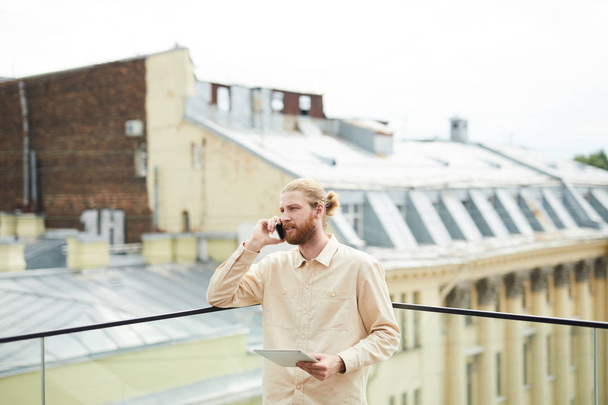 Young bearded man looking at digital tablet and consulting somebody on mobile phone while standing outdoor on the roof of building  - Foto, afbeelding
