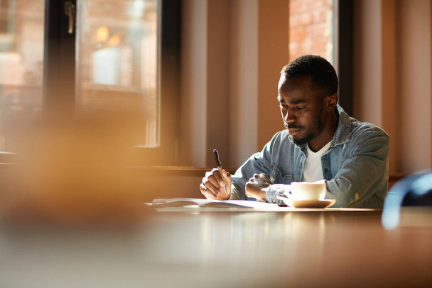 Düşünceli Afrikalı bir adam elinde bir fincan kahveyle oturmuş not defterine not alıyor ve kafede yeni fikirler düşünüyor. - Fotoğraf, Görsel
