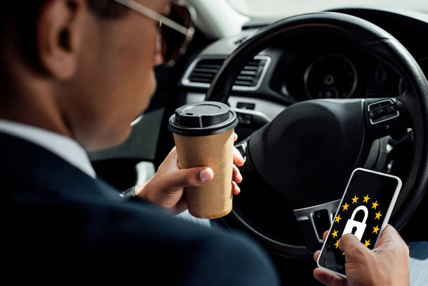 back view of african american businessman using smartphone and drinking coffee in car with gdpr illustration - Фото, изображение