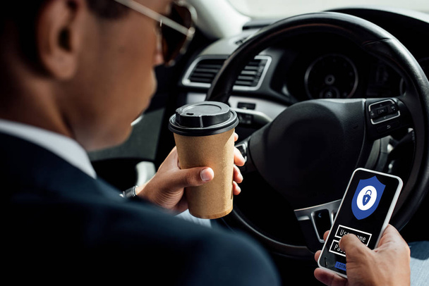 back view of african american businessman using smartphone with secure access illustration and drinking coffee in car - Fotó, kép